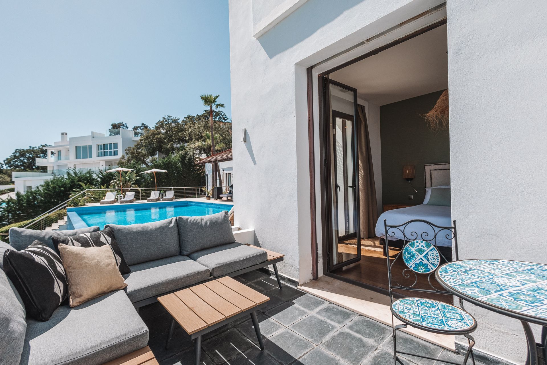 A white villa balcony with a pool view, featuring a sofa, table, and access to a bedroom with a bed.
