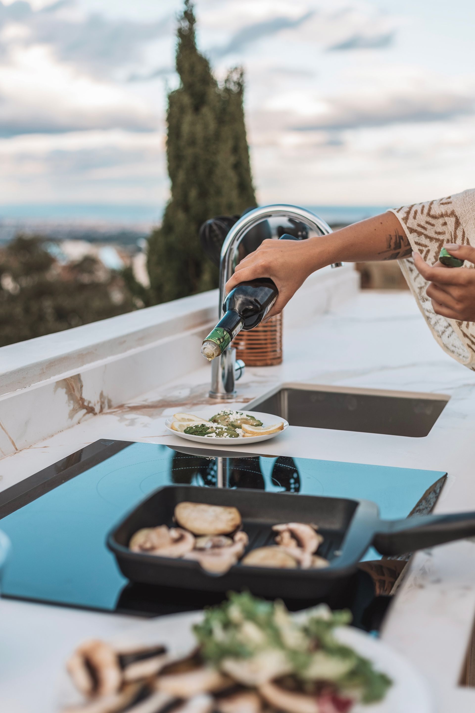 Person cooking outdoors, pouring liquid from bottle into a pan with mushrooms. The background includes a sink, and scenic views.