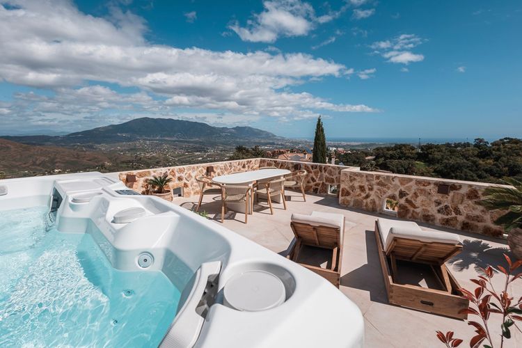 Hot tub on a sunny patio with lounge chairs, a dining table, and a scenic view of a town and mountains.