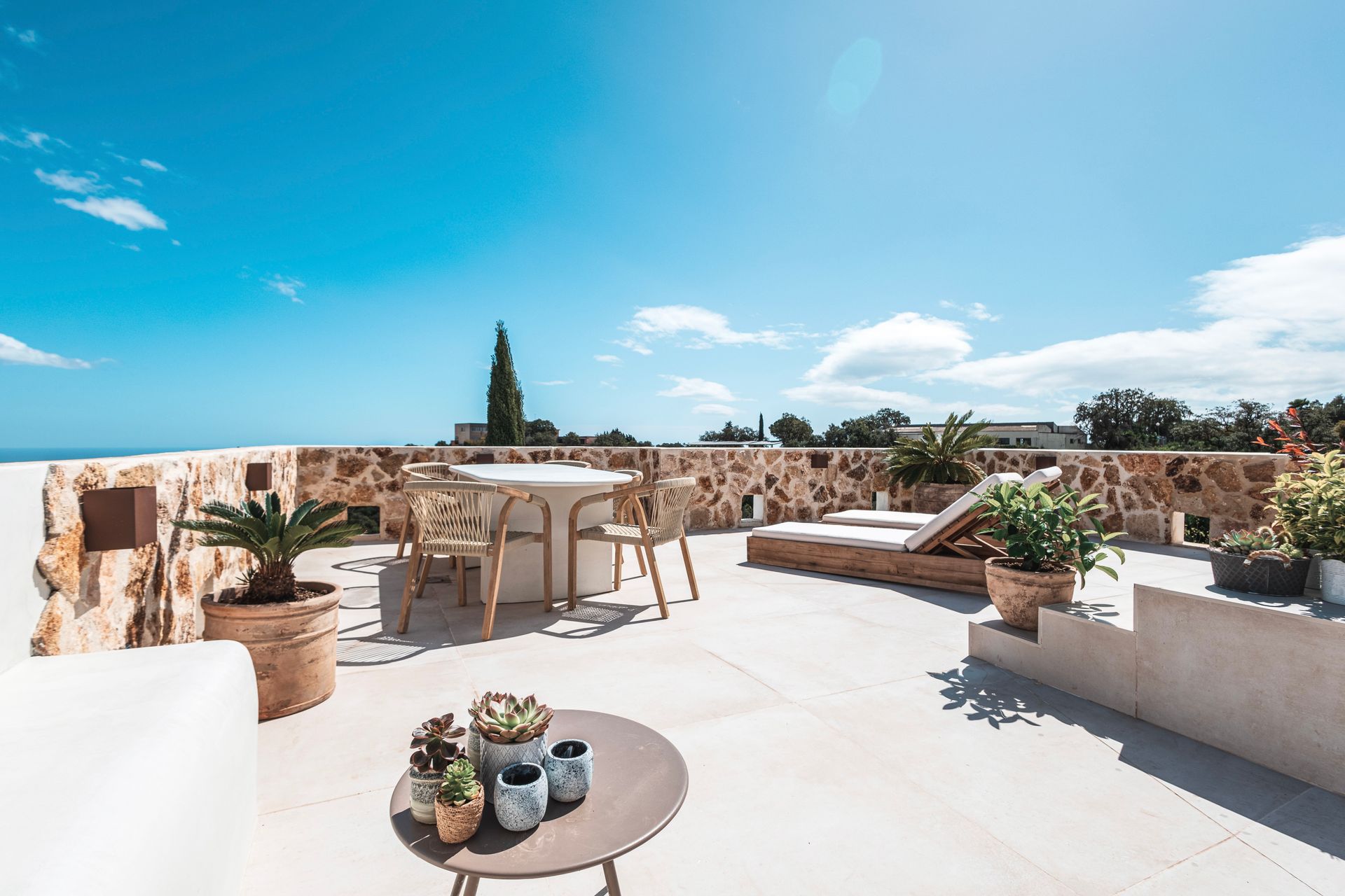 Rooftop terrace with stone walls, table and chairs, lounge chair, potted plants, and a blue sky.