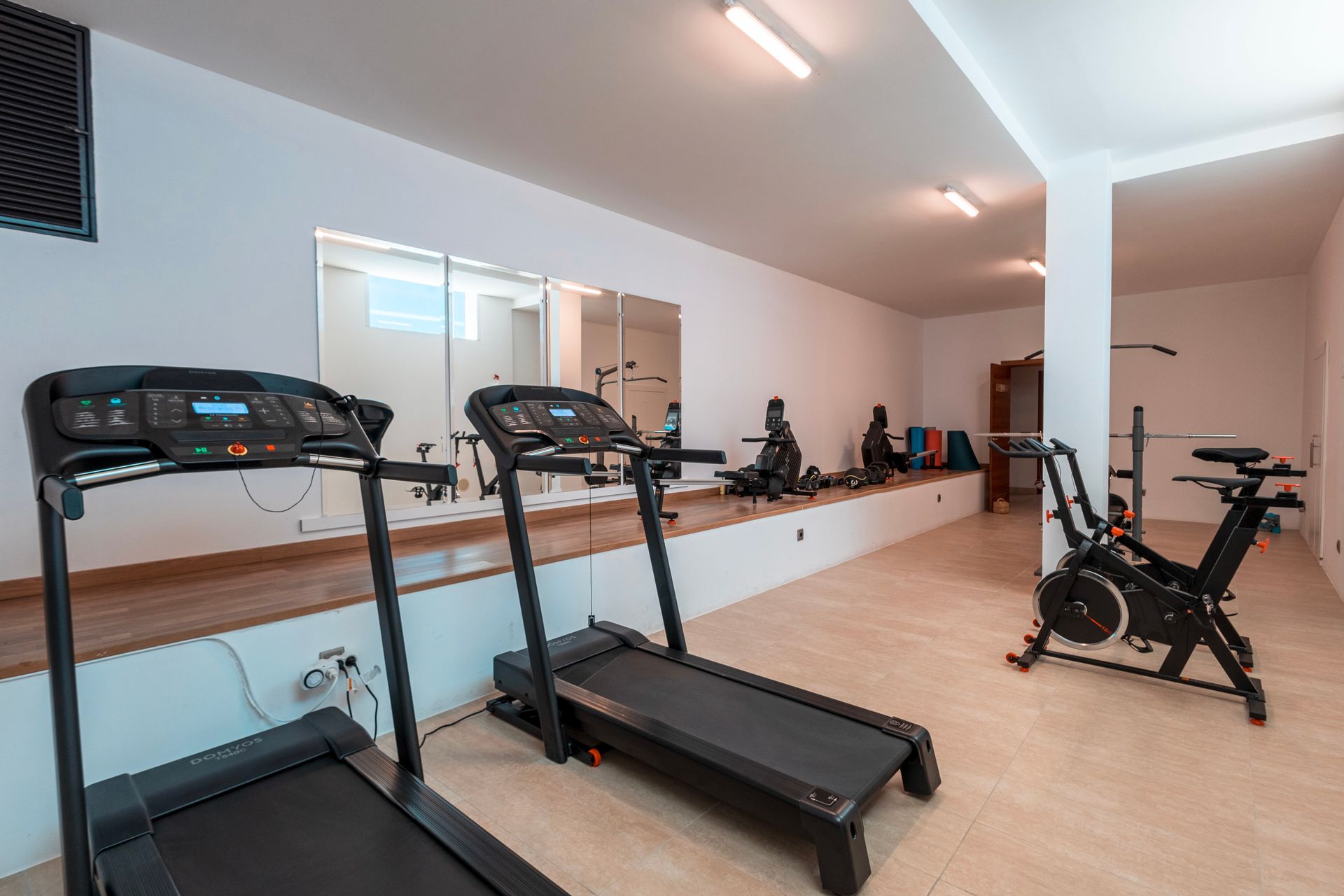A home gym with two treadmills, an exercise bike, and mirrors on the wall. White walls and bright lighting.