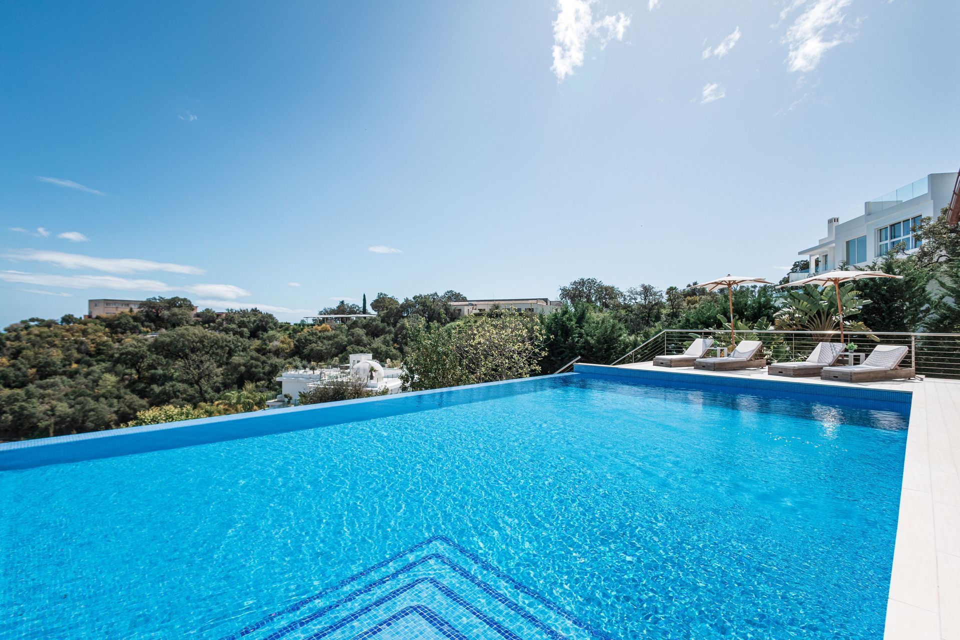 Infinity pool overlooking a landscape with blue sky. Lounge chairs sit by the pool, and a white building is visible in the distance.