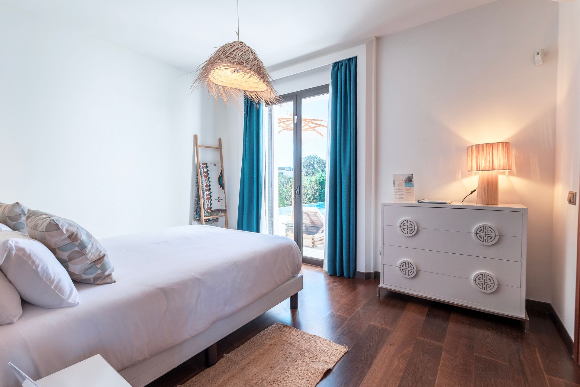 Bright bedroom with a white bed, dresser, and walls. Teal curtains frame a view of a pool.