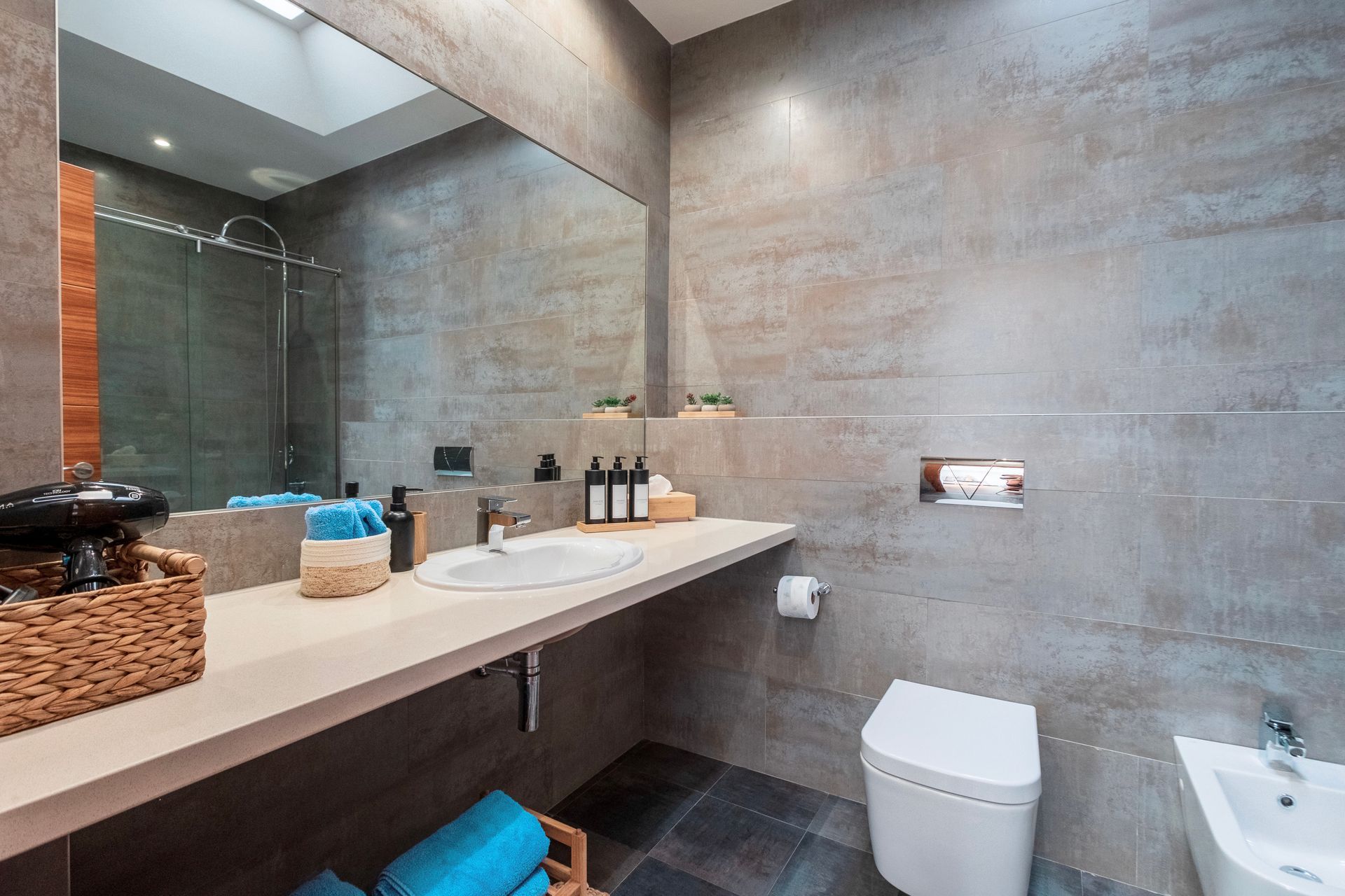 Modern bathroom with a large mirror, floating vanity, and a toilet. Gray tiled walls and a skylight provide natural light.