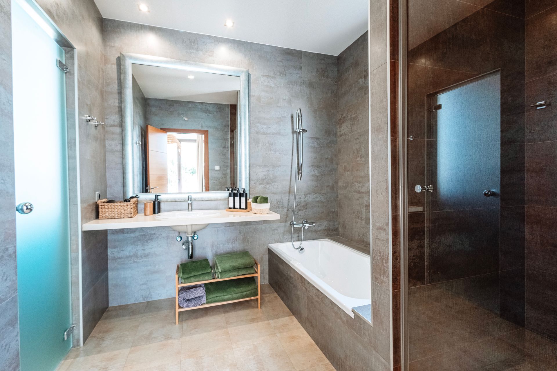 Modern bathroom with gray tiled walls, a rectangular mirror, and a bathtub. Includes a shower, sink, and frosted glass door.