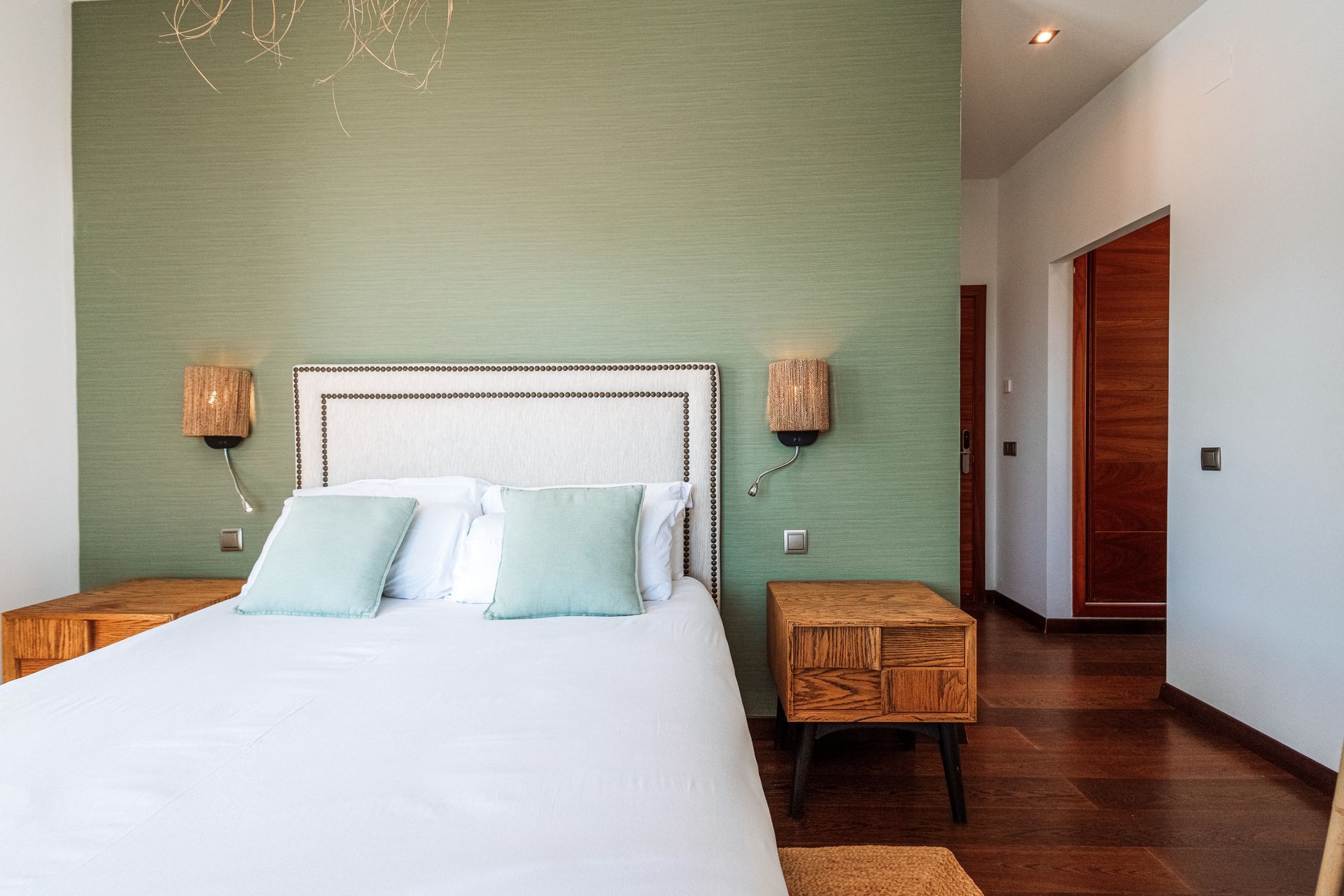 Bedroom with a bed, green accent wall, wooden nightstands, and a hallway with a closed wooden door.