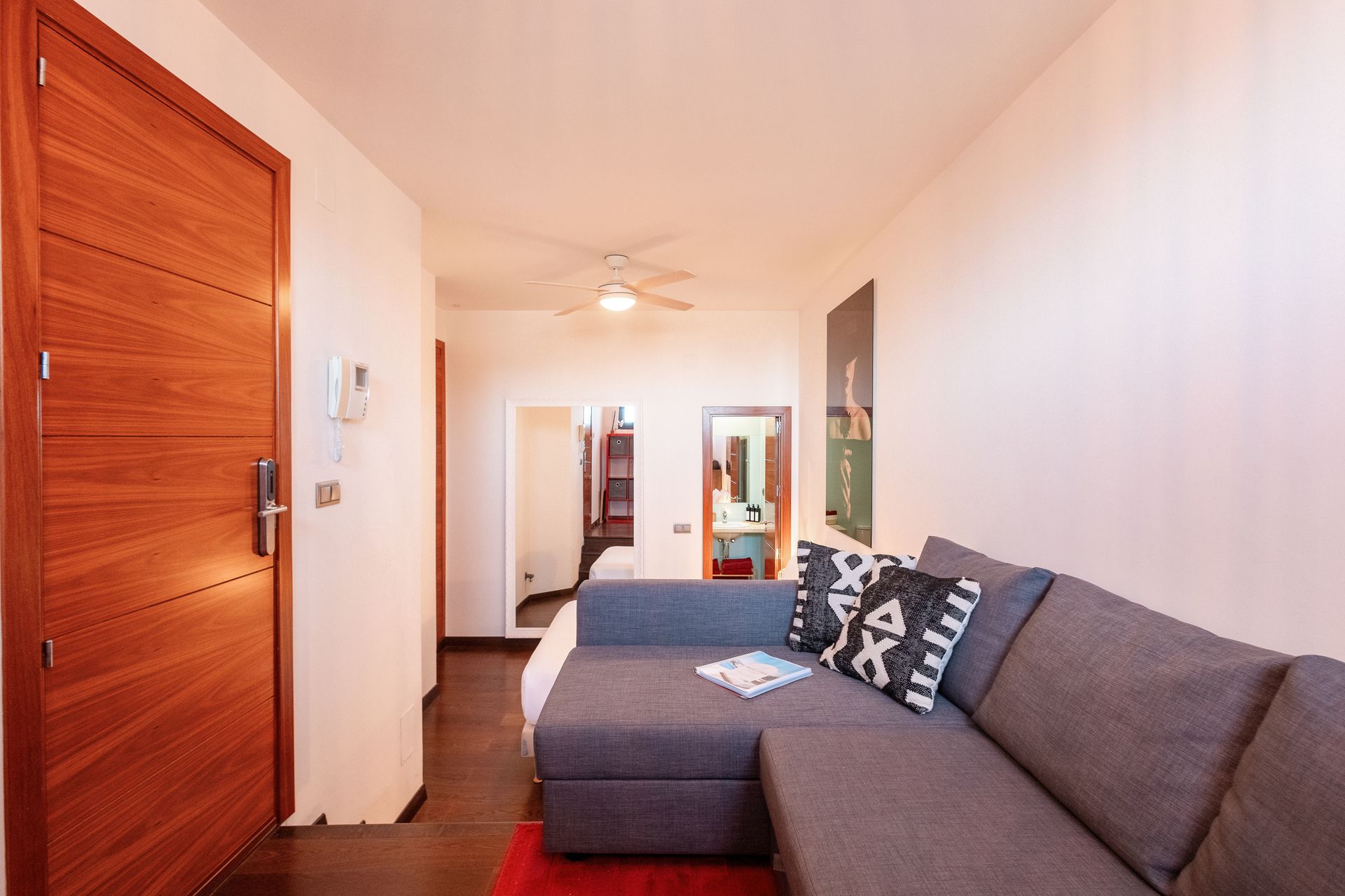 A small apartment interior with a gray sectional sofa, red rug, and wooden door leading to a bedroom and bathroom.
