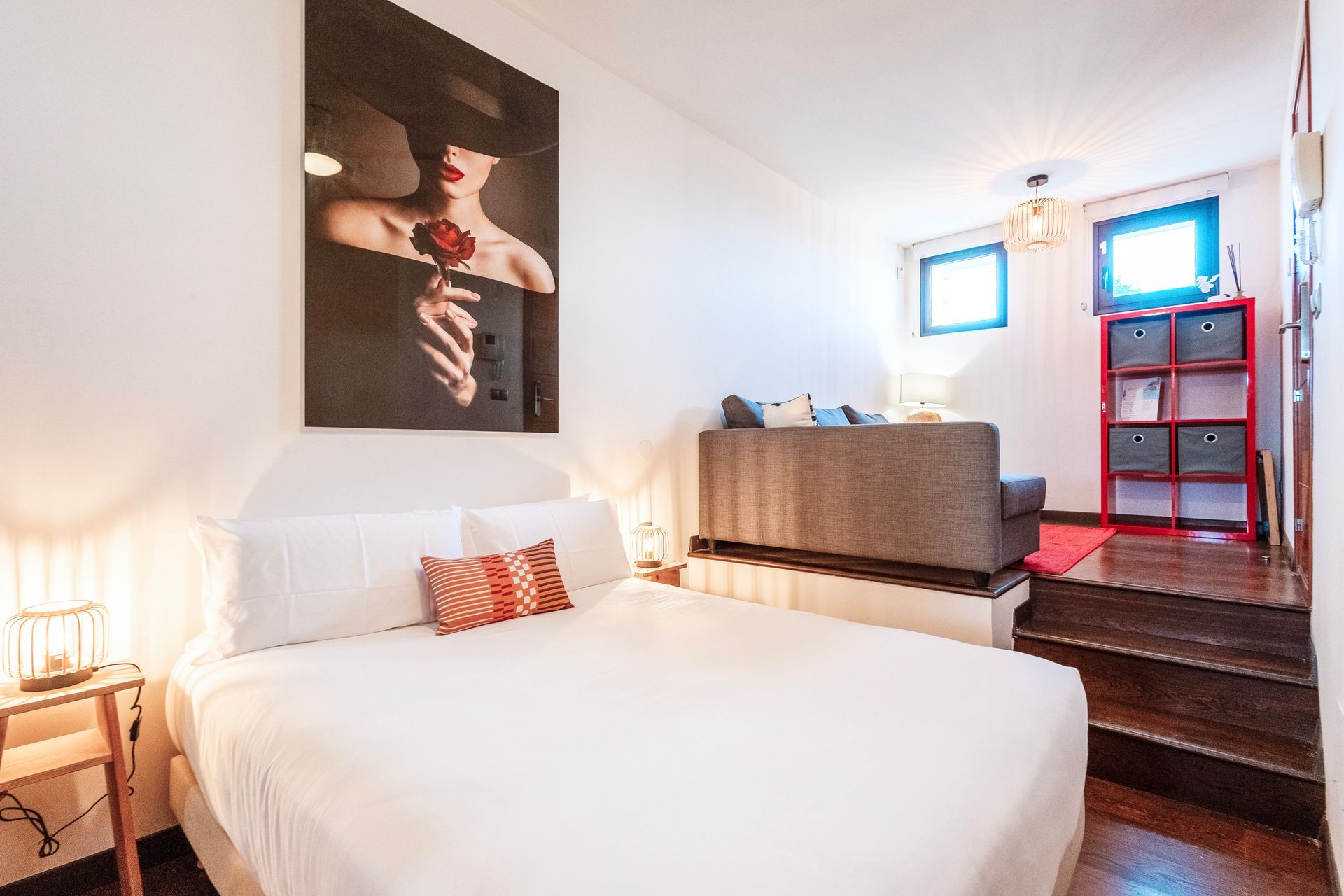 Bedroom with a raised living area; white bedding, artwork, sofa, red shelving unit, dark wood steps.