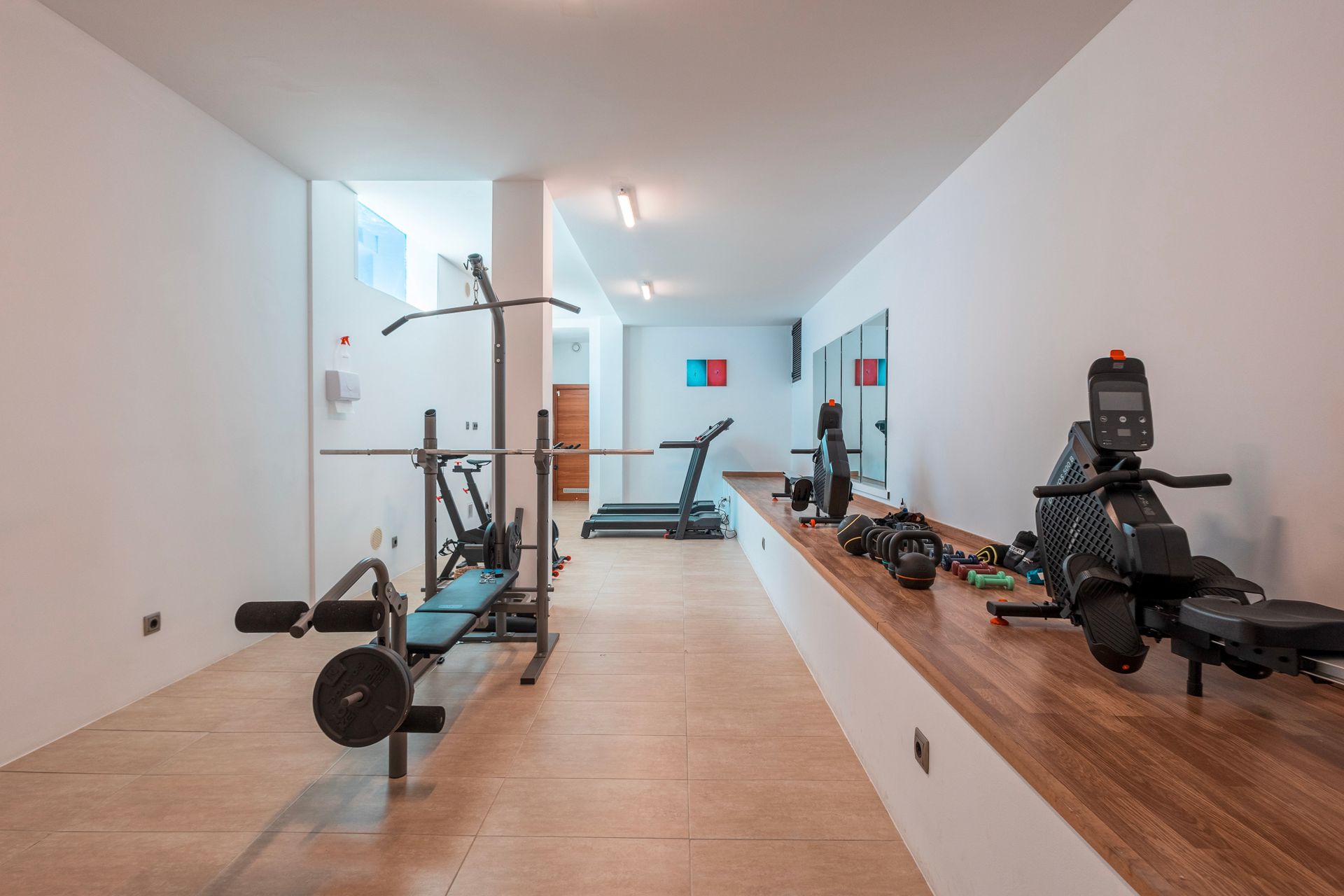 Gym with workout equipment including a bench, pull-up bar, treadmill, and rowing machine, located indoors with white walls.