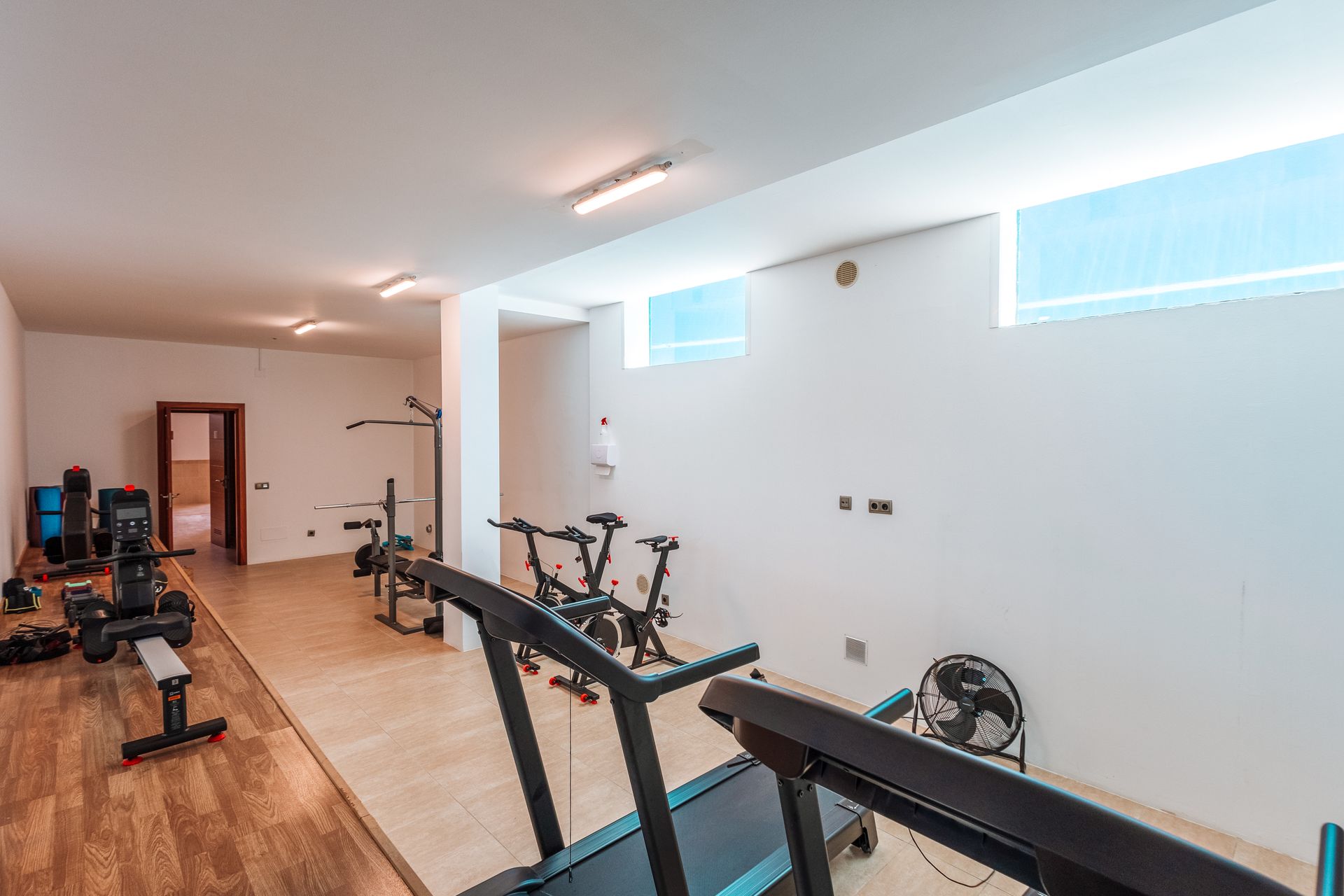 Home gym with various exercise machines, including a treadmill, stationary bikes, and weight machines. The room is white with light wood floors.