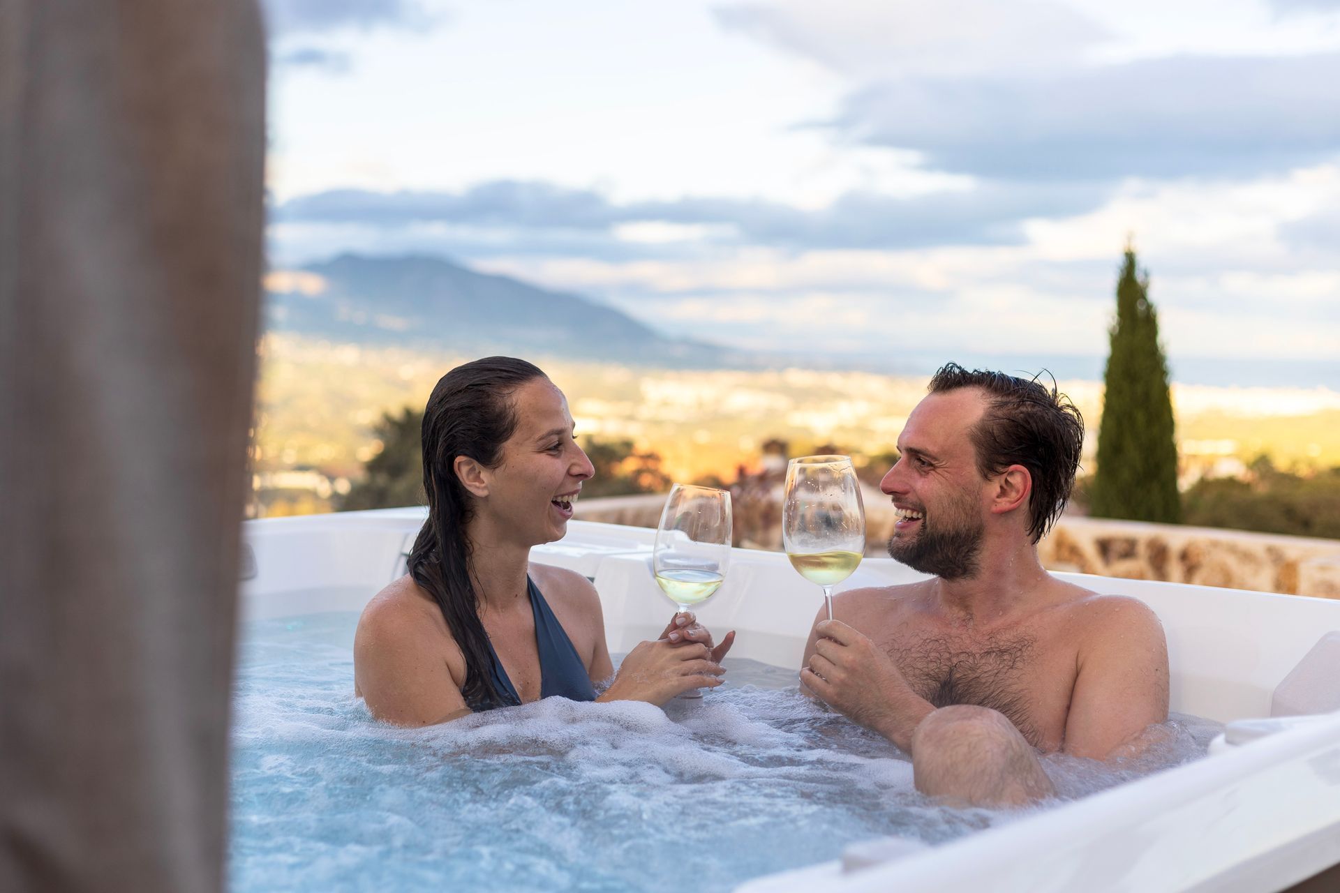 A couple in a hot tub, drinking white wine, with a scenic mountain view in the background.