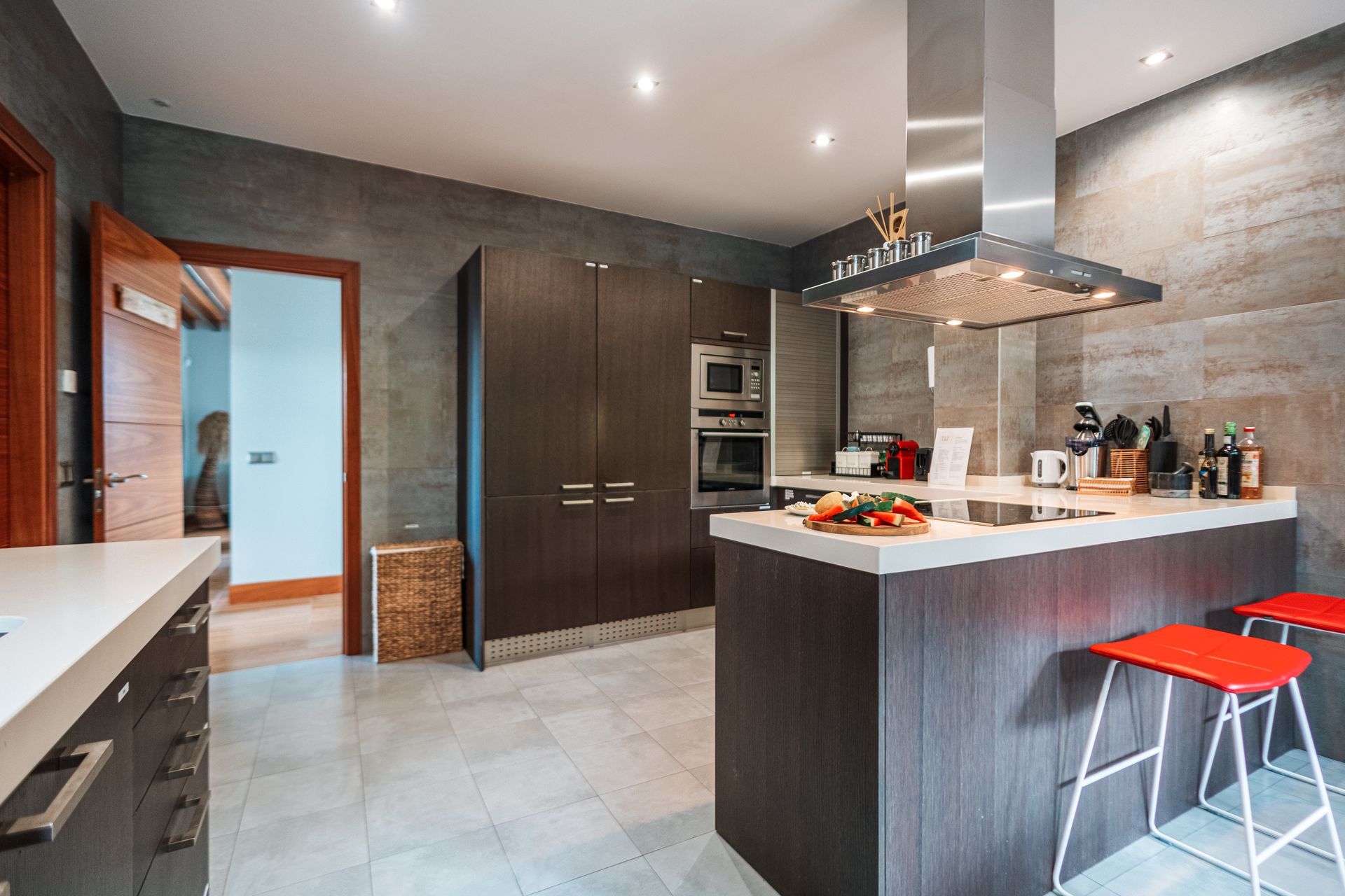 Modern kitchen with a dark wood island and cabinets, stainless steel range hood, and gray tile walls. Two red stools sit at the island.