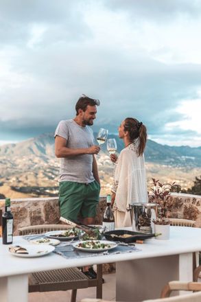 Couple toasting with wine at an outdoor dinner overlooking mountains. Man smiles at woman as they hold up glasses.
