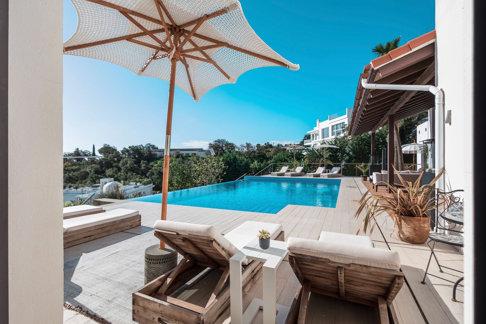 Poolside scene with a pool, lounge chairs, and an umbrella on a sunny day. White buildings and lush greenery are visible in the background.