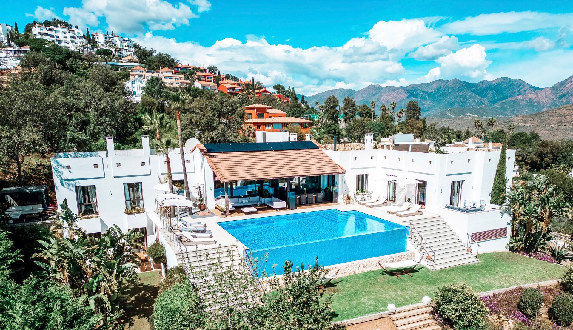 White villa with a blue pool, nestled in a green landscape under a cloudy sky, with mountains in the background.