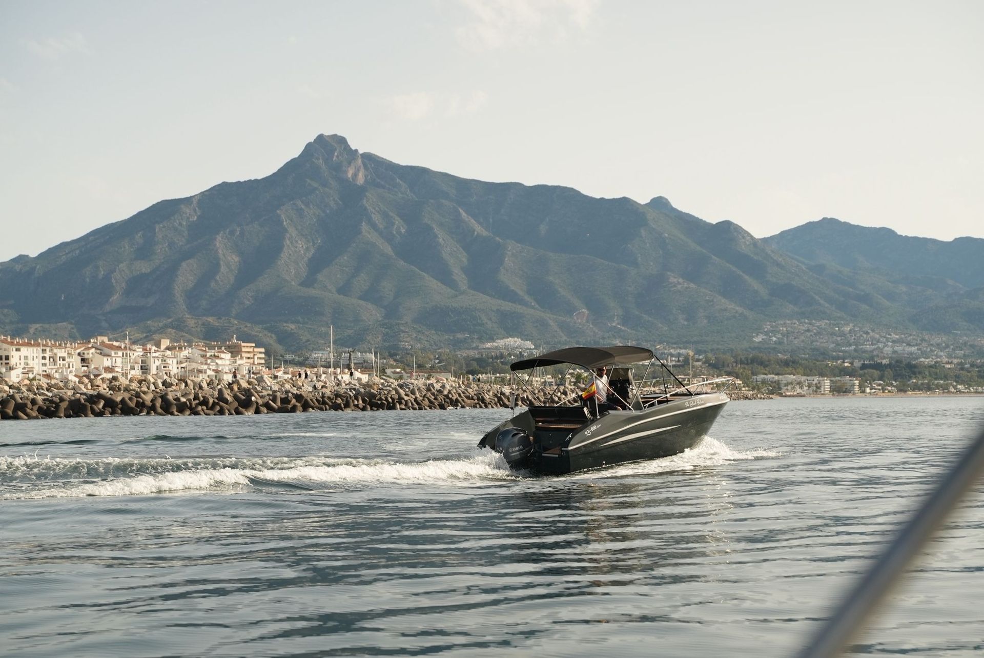 A black speedboat on water, heading towards a mountain range, with a town on the left.