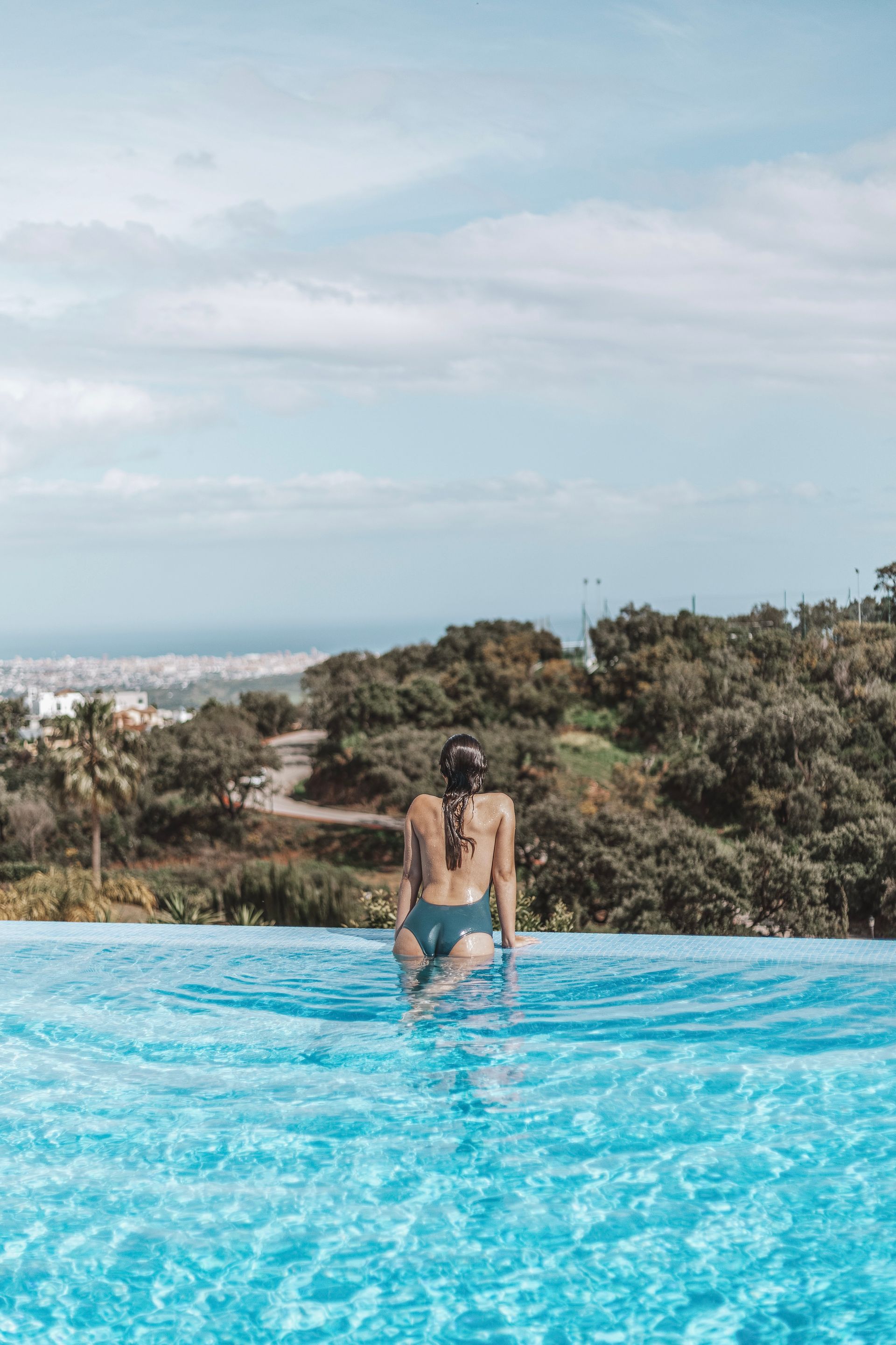 Woman in a swimsuit standing at the edge of an infinity pool, overlooking a city and green hills under a cloudy sky.