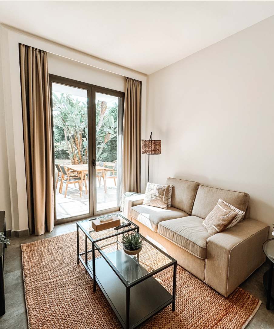 A living room with a tan couch, rug, and glass coffee table, opening onto a patio. Brown curtains frame a glass door.