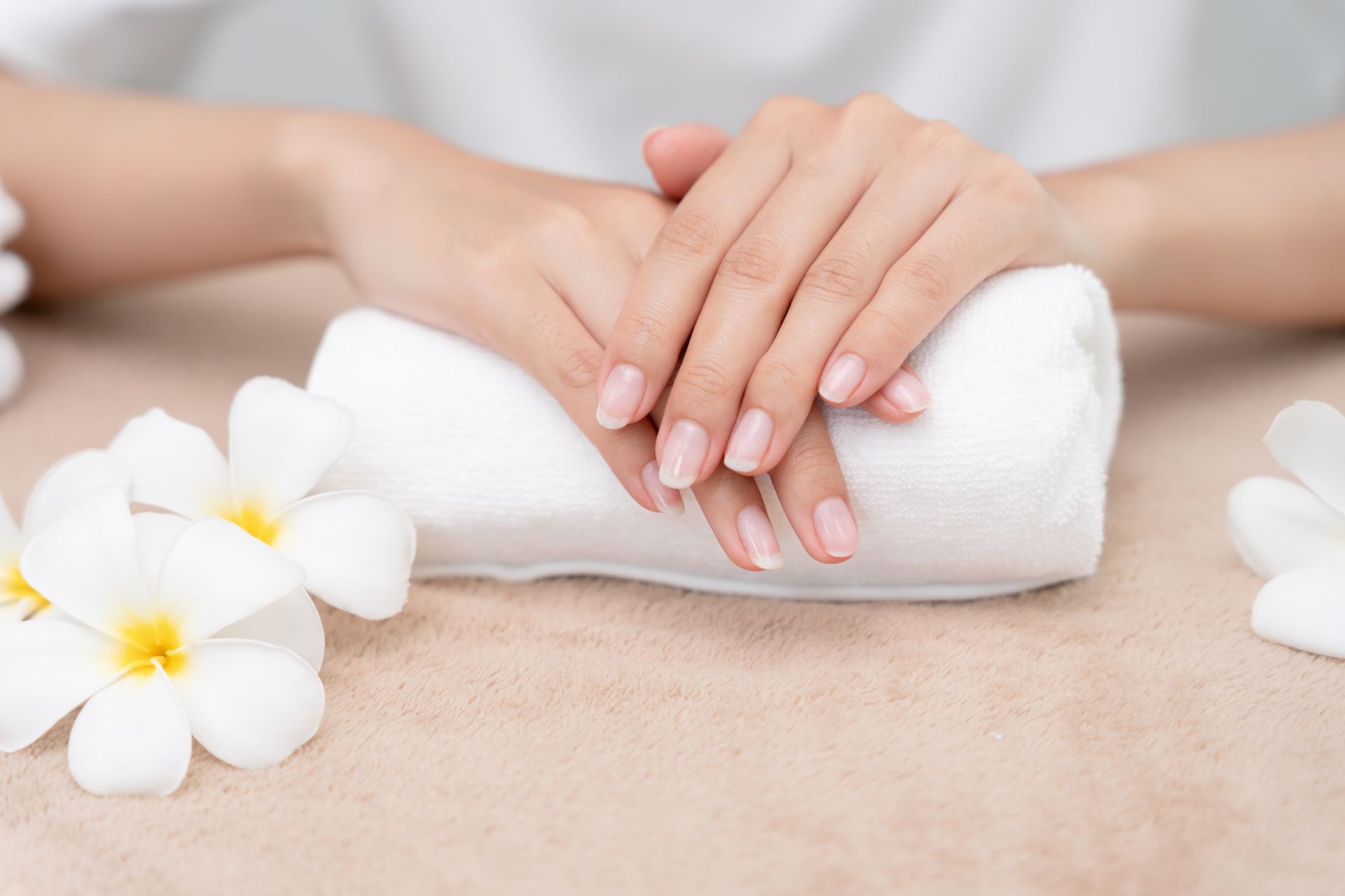 Hands with manicured nails resting on a rolled white towel, surrounded by white flowers, neutral background.