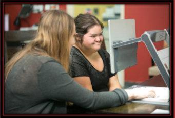 two women are sitting at a desk looking at a computer screen