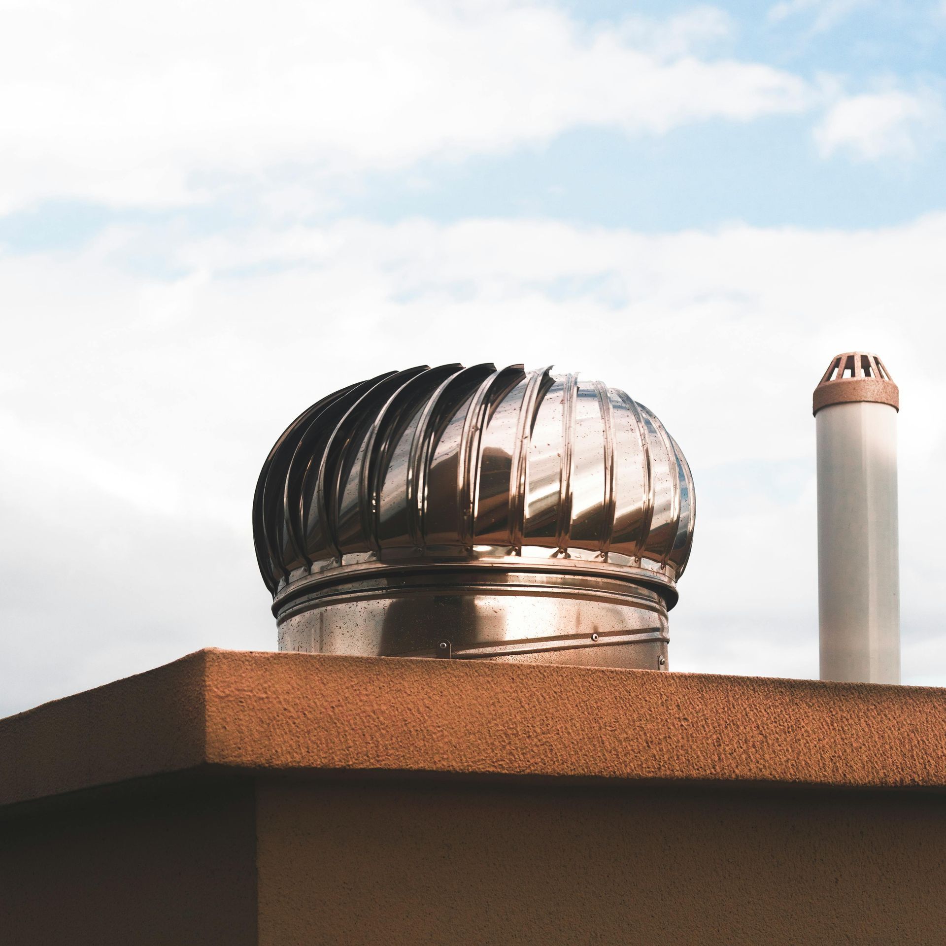 Shiny metal roof vent and a cylindrical chimney against a cloudy sky.