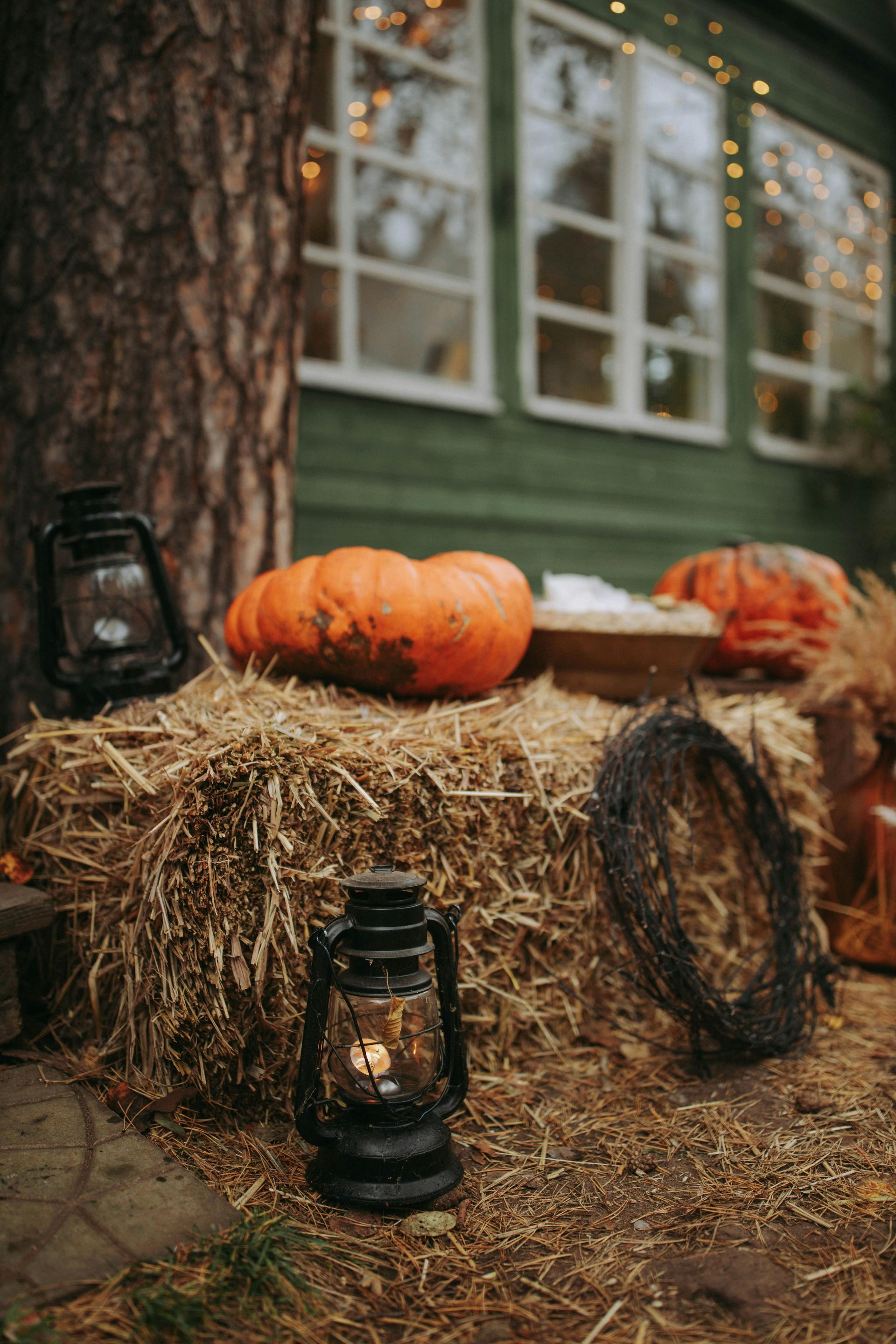 Hay bales with pumpkins and lanterns in front of a green house with string lights.