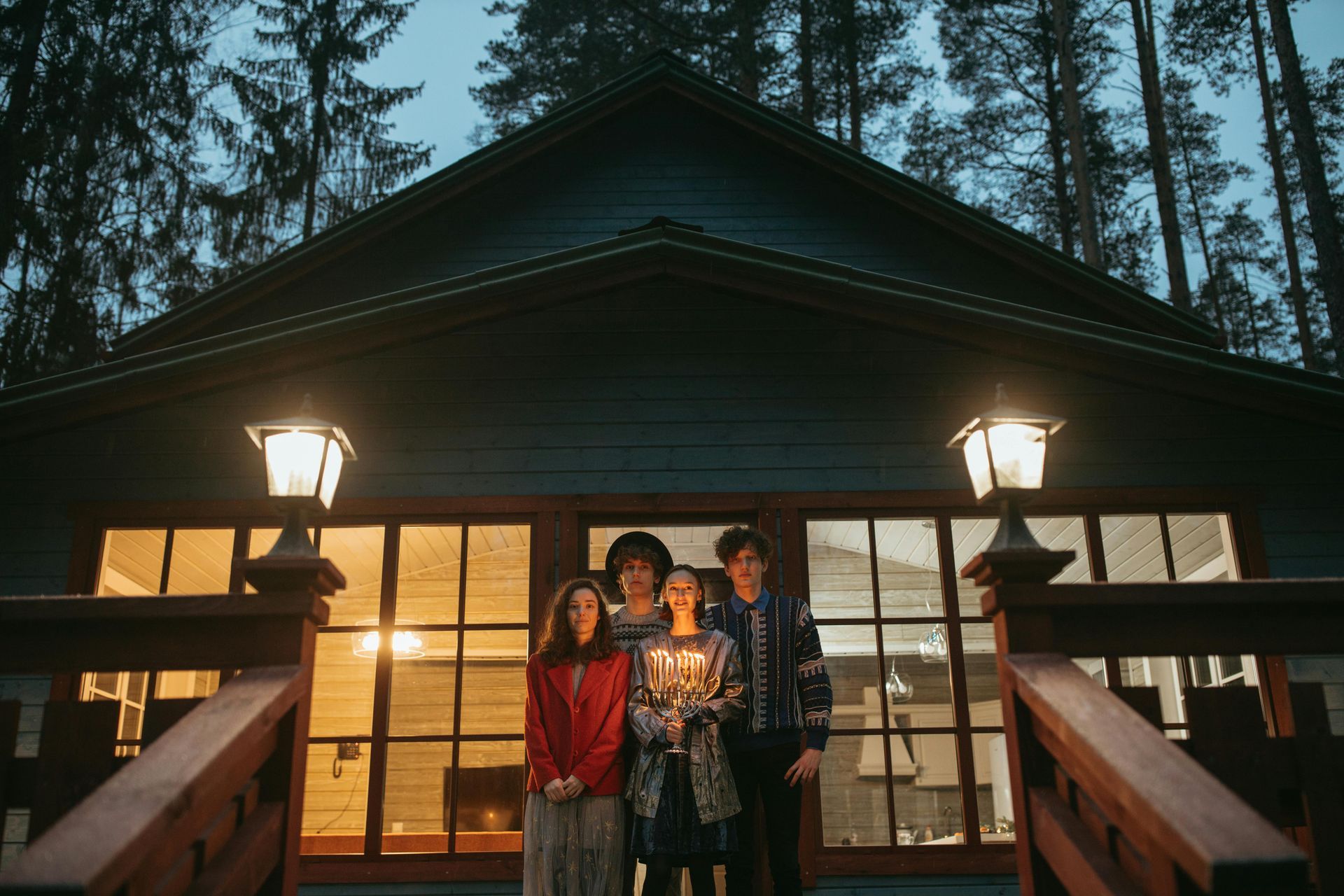 Four people standing on porch of a blue cabin, lit by outdoor lamps. Dusk setting, surrounded by trees.