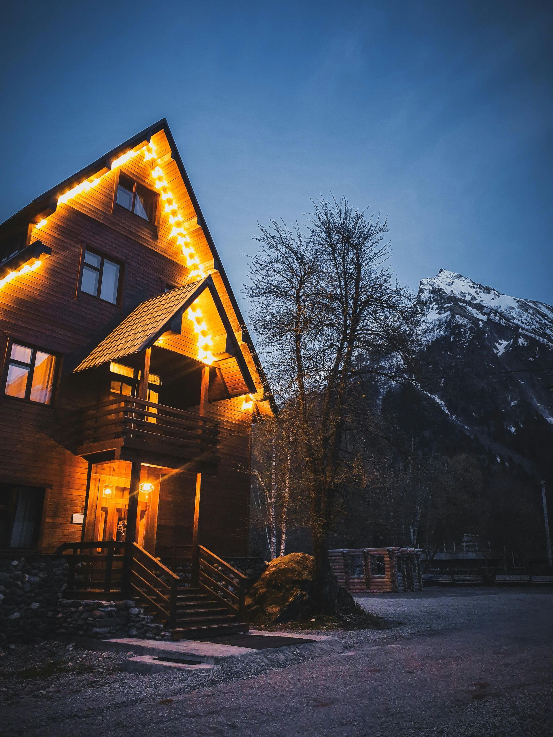 Wooden lodge lit with warm lights at dusk, with mountain in background.