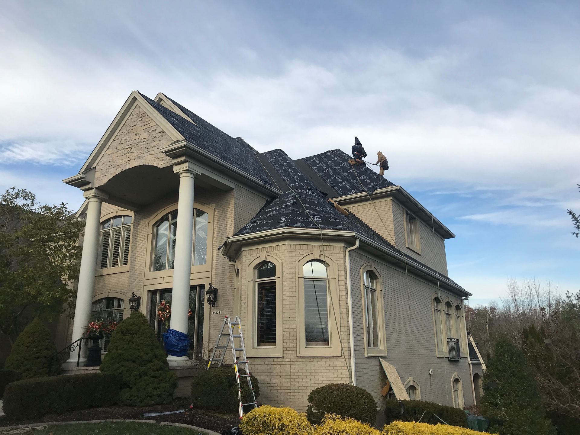 Two-story house with workers on the roof; cream and brick exterior, large columns, blue sky.