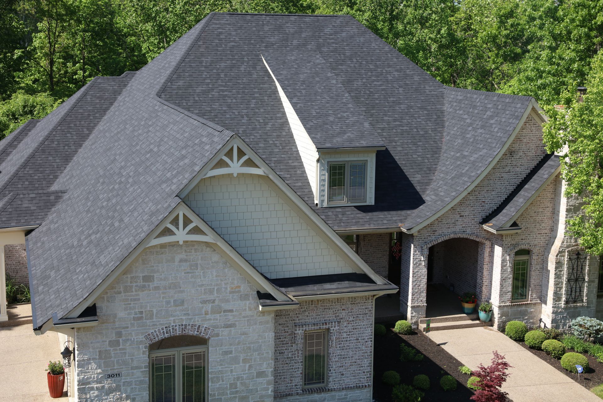 House with dark gray roof, light brick facade, and lush green trees.