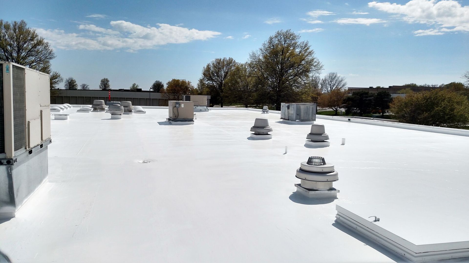 The roof of a building with a white coated roof and trees in the background.