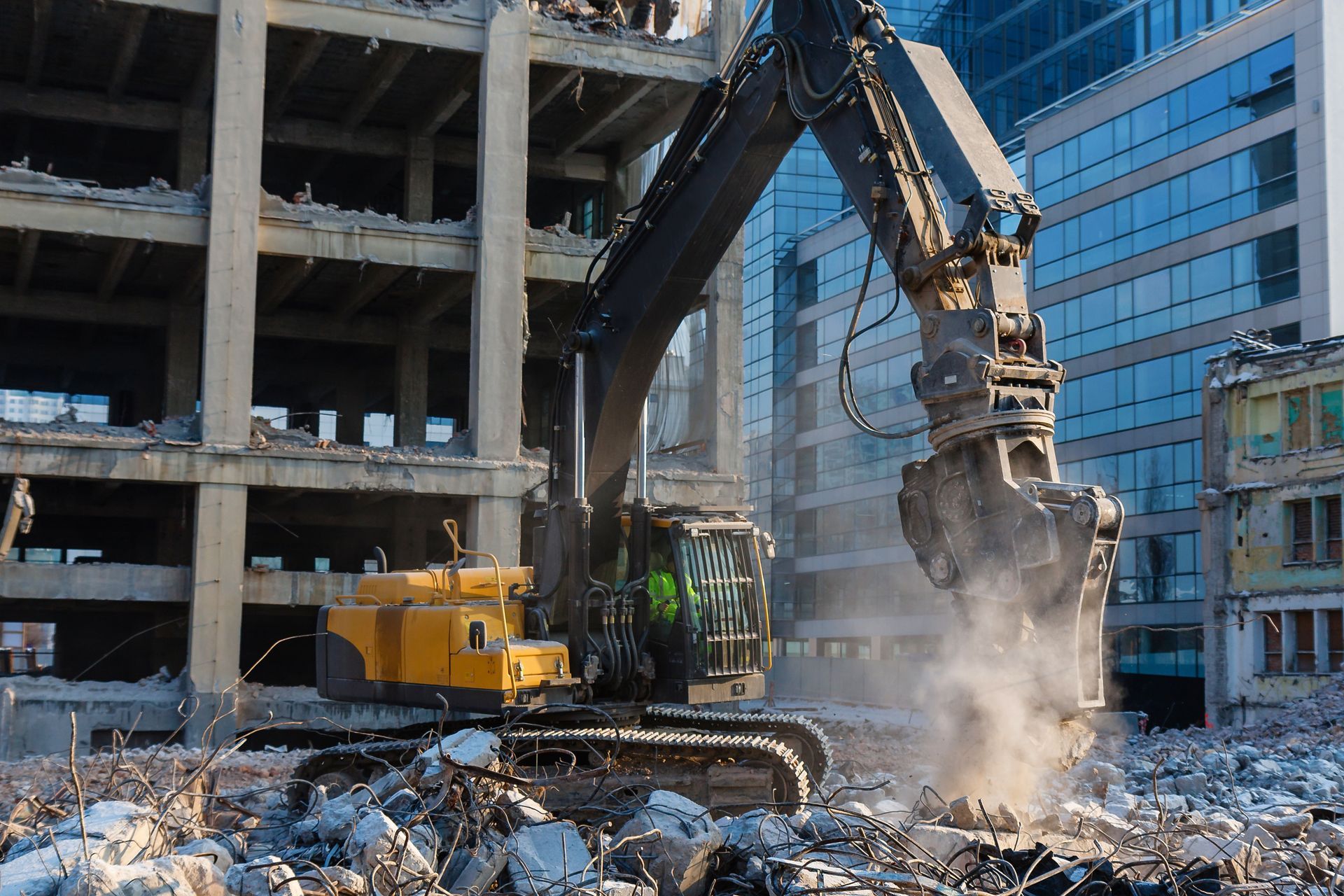 Yellow excavator demolishing a building, with dust and rubble in an urban setting.