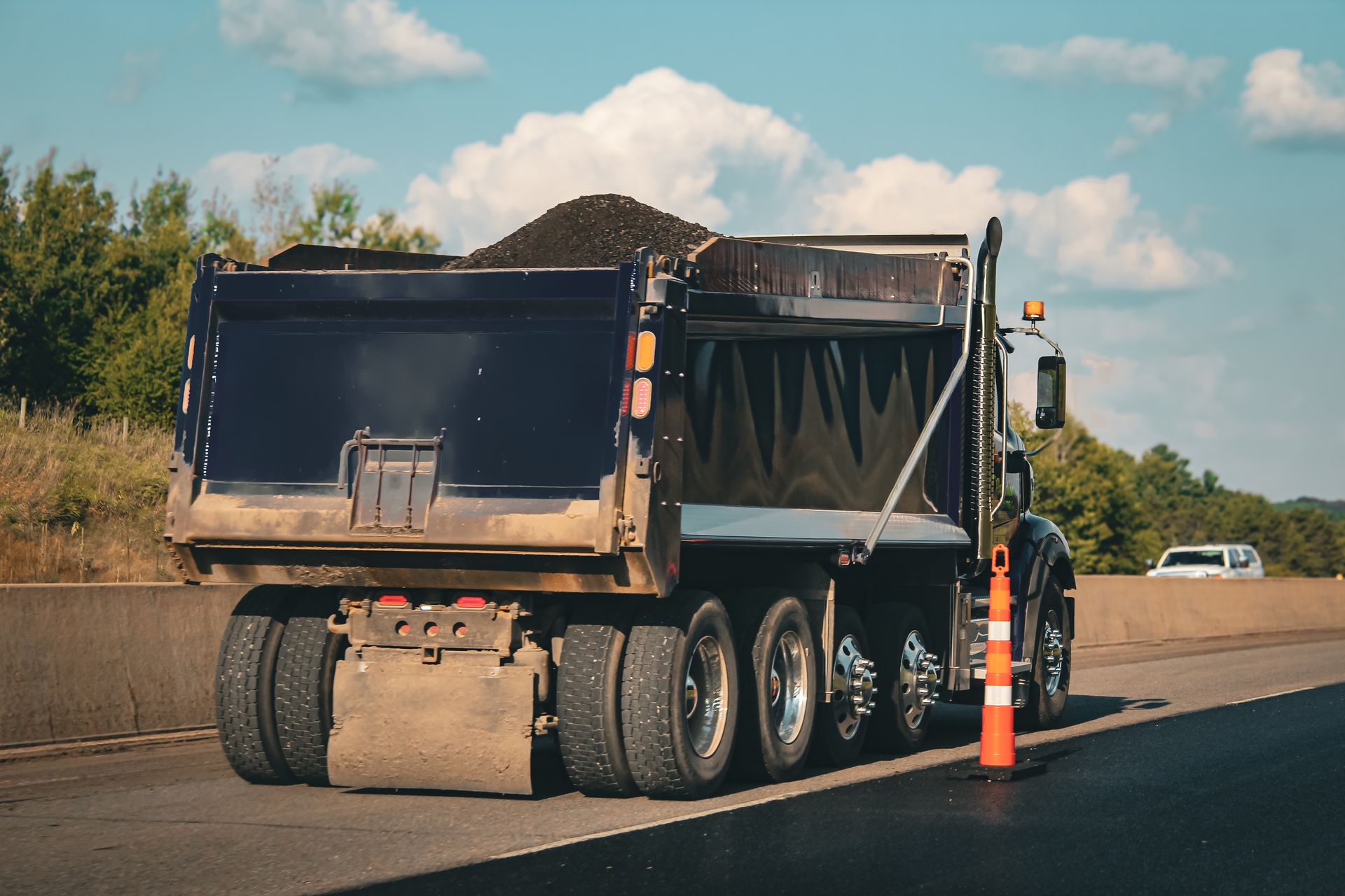 Rear view of a dump truck loaded with dark asphalt on a highway next to a traffic cone.