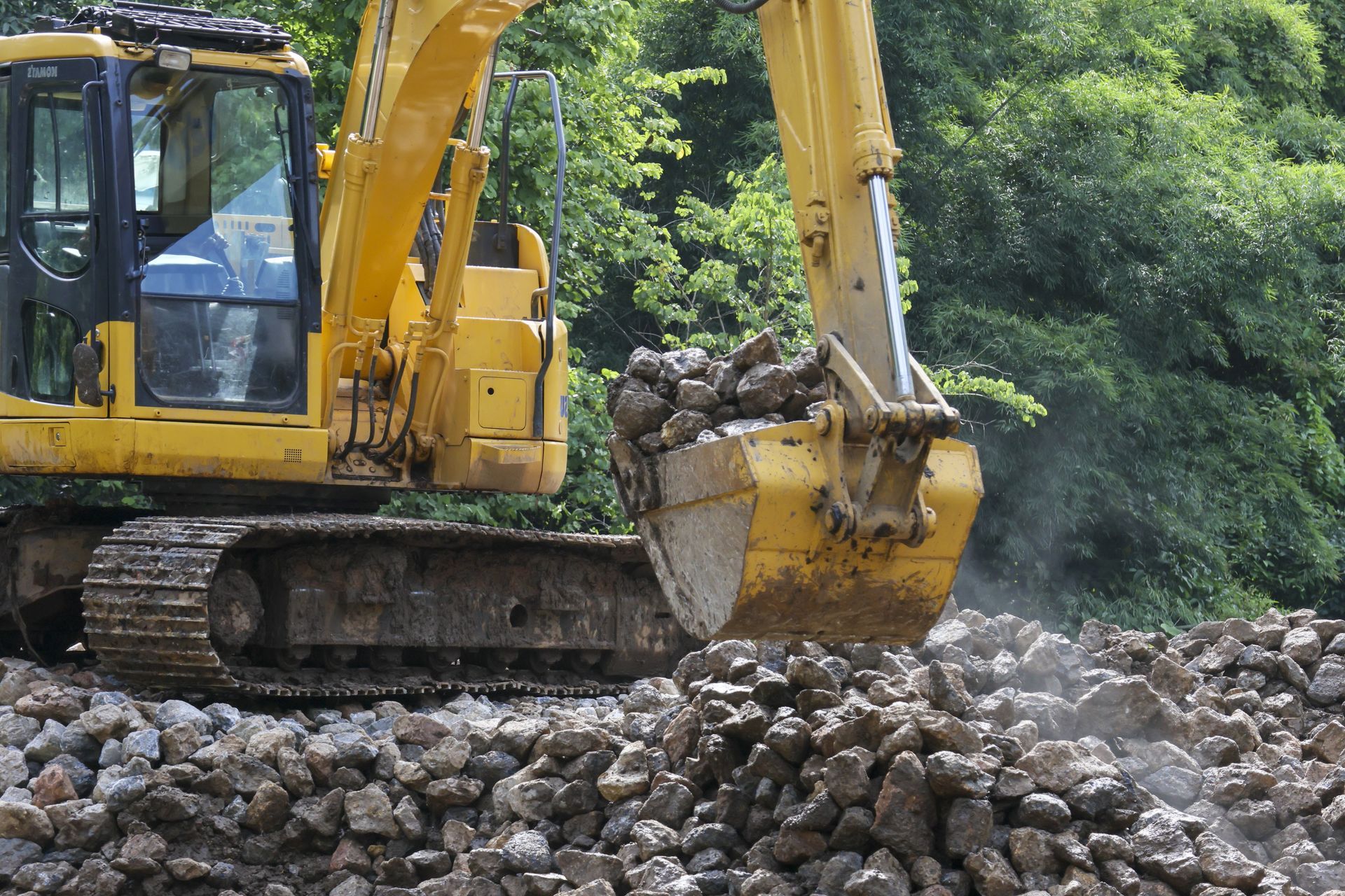 Yellow excavator scoops rocks at a construction site, trees in background.