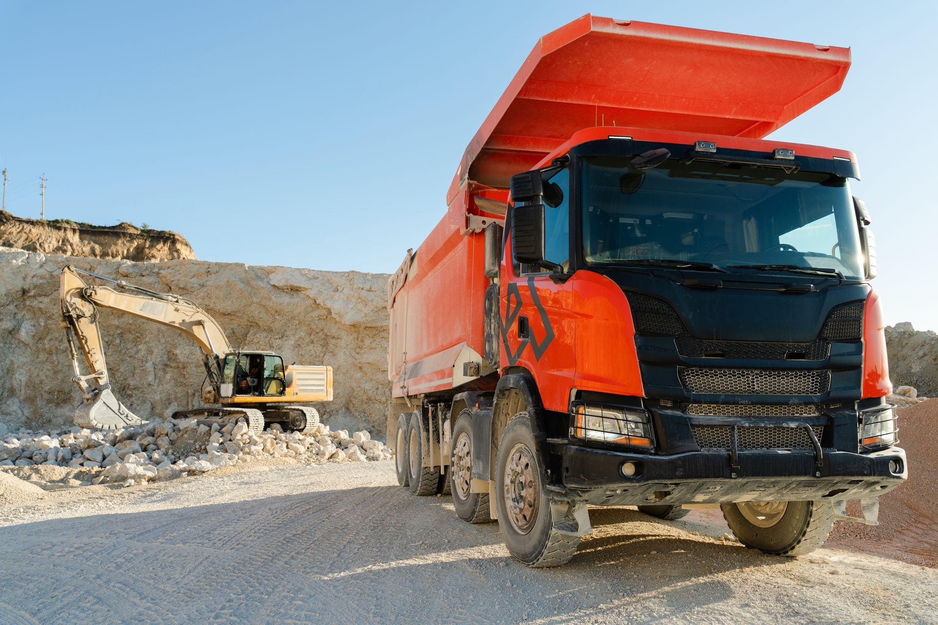 Orange dump truck parked at a quarry, excavator in the background, blue sky.