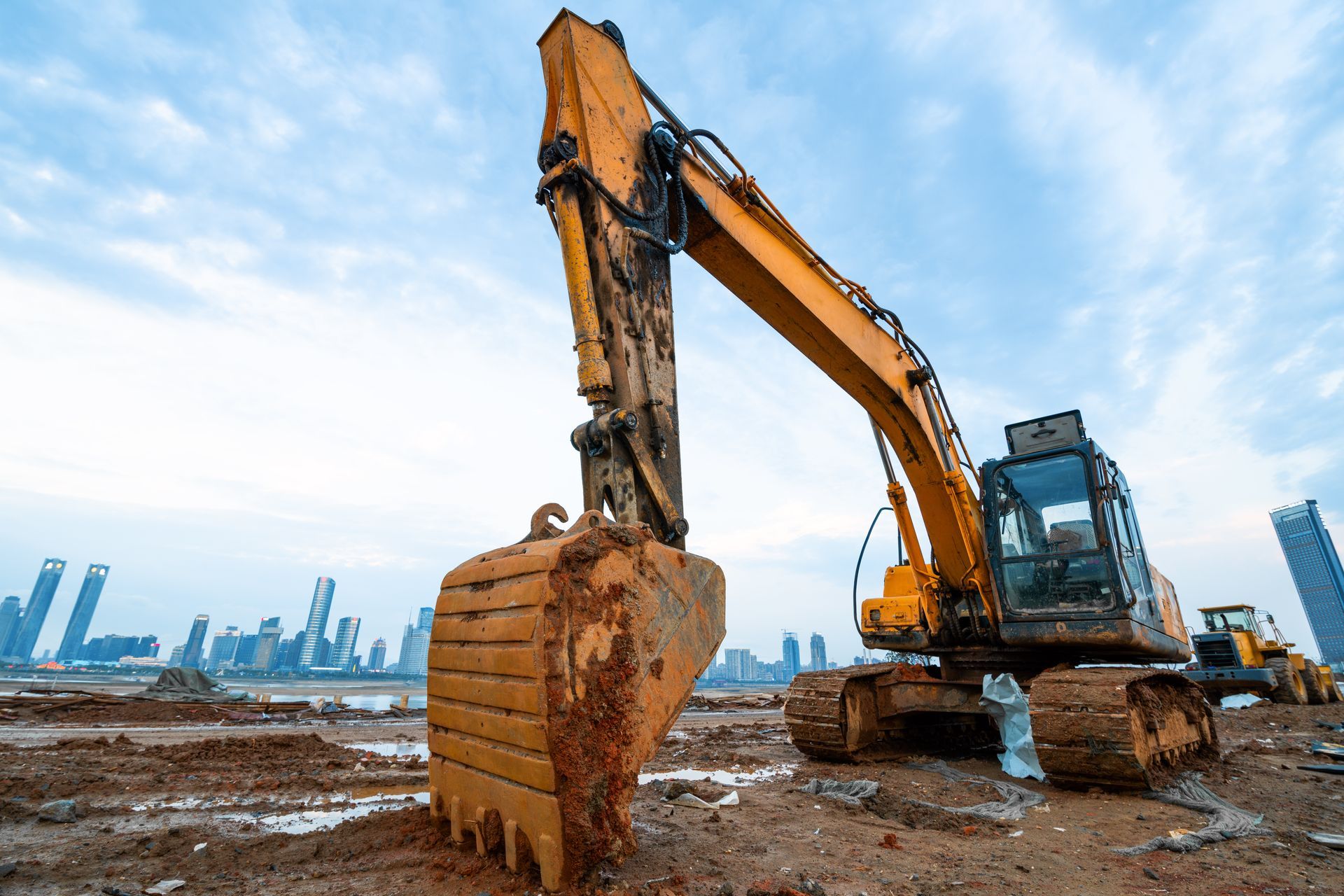Yellow excavator on muddy construction site, city skyline in the background.