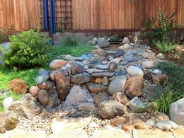 A dry creek bed made of various sized gray and brown rocks, bordered by lush green plants and a wooden fence.