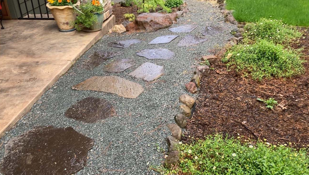 A stone pathway of uneven, flat stepping stones set into grey gravel, bordered by mulch and greenery in a residential yard.