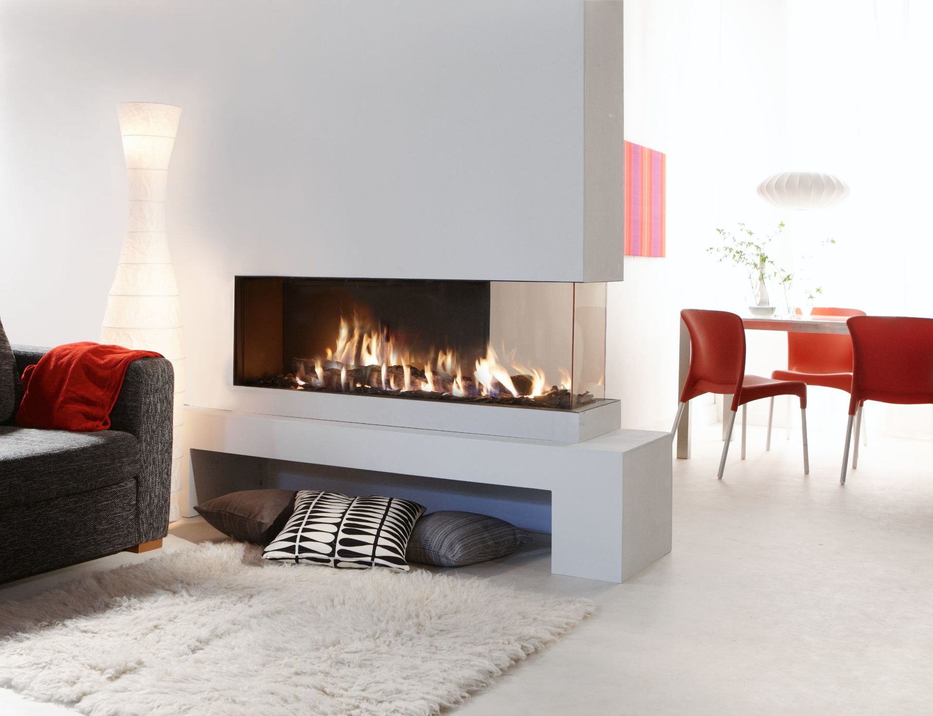 Modern white fireplace with flames visible through glass. A fluffy white rug and dark gray sofa with red throw and pillows are in front of the fireplace. The room is bright and airy.