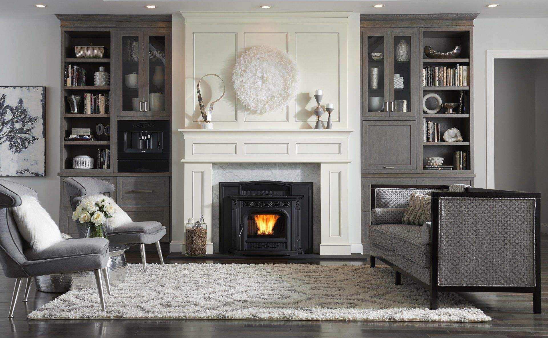 Living room with fireplace, built-in bookshelves, two gray chairs, and a gray sofa. A lit fireplace sits below a white mantle.