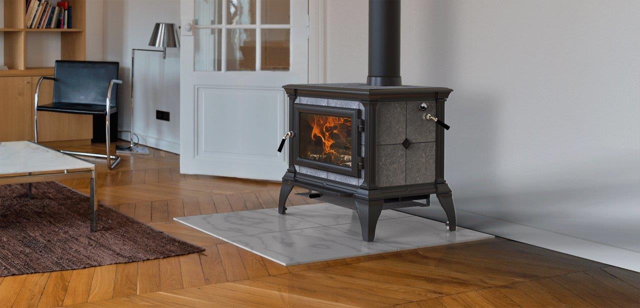 A wood-burning stove with a visible fire sits on a tile hearth in a light-filled room. The floor is hardwood.