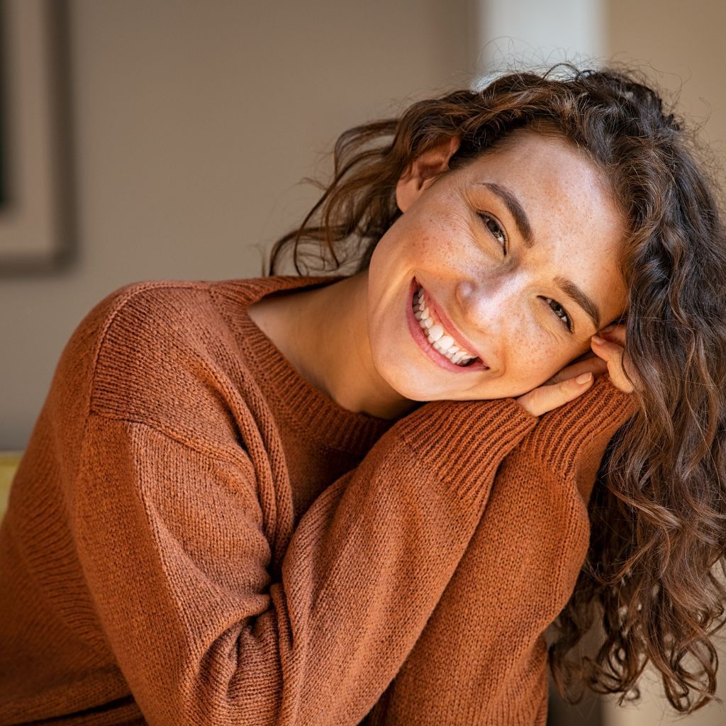 Woman with curly hair, freckles, and a warm smile, wearing an orange sweater, leaning on her arms.