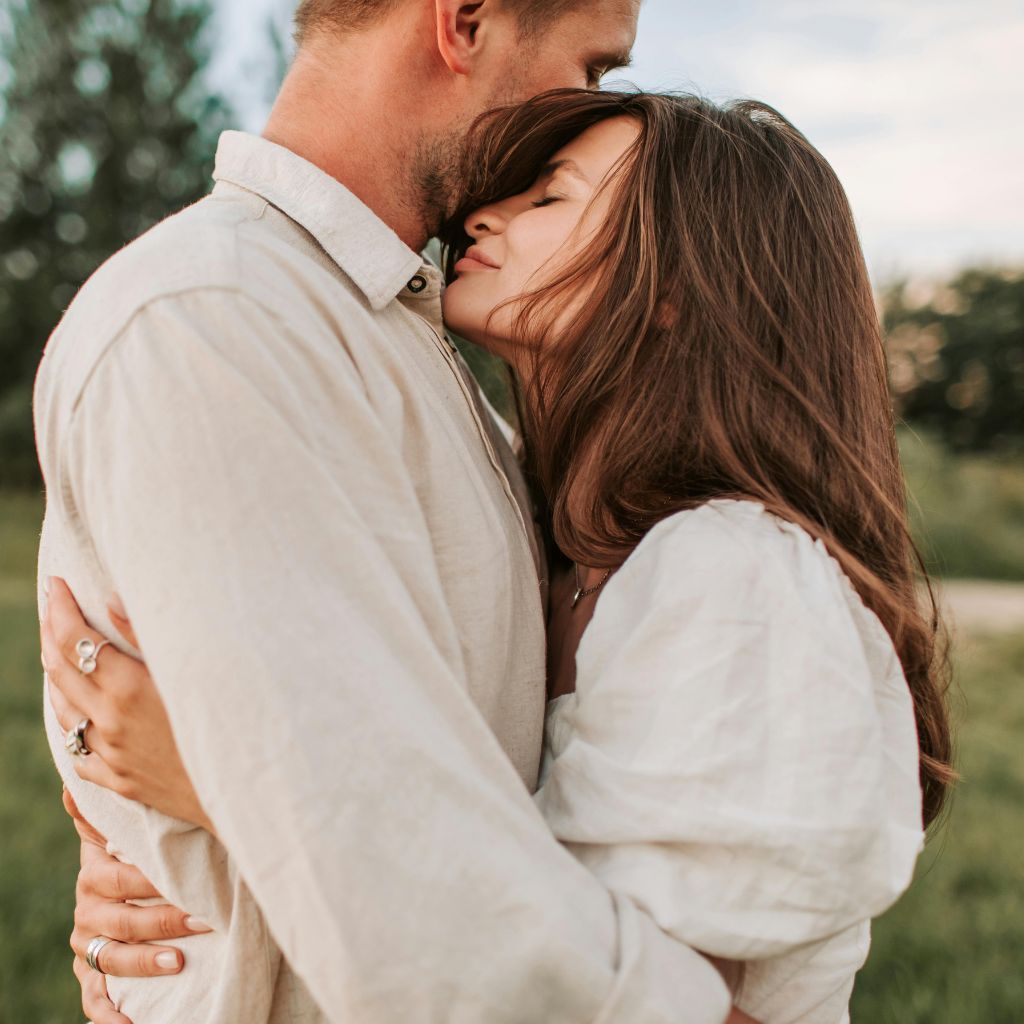 Couple embraces outdoors, woman resting head on man's chest. Soft, neutral tones, natural light.