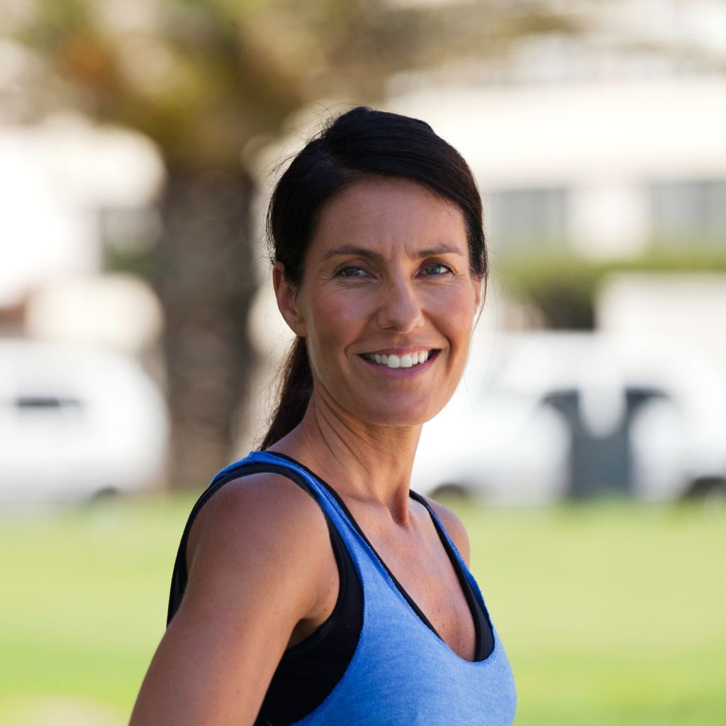 Woman in blue athletic top smiles outdoors, blurred background of greenery and buildings.