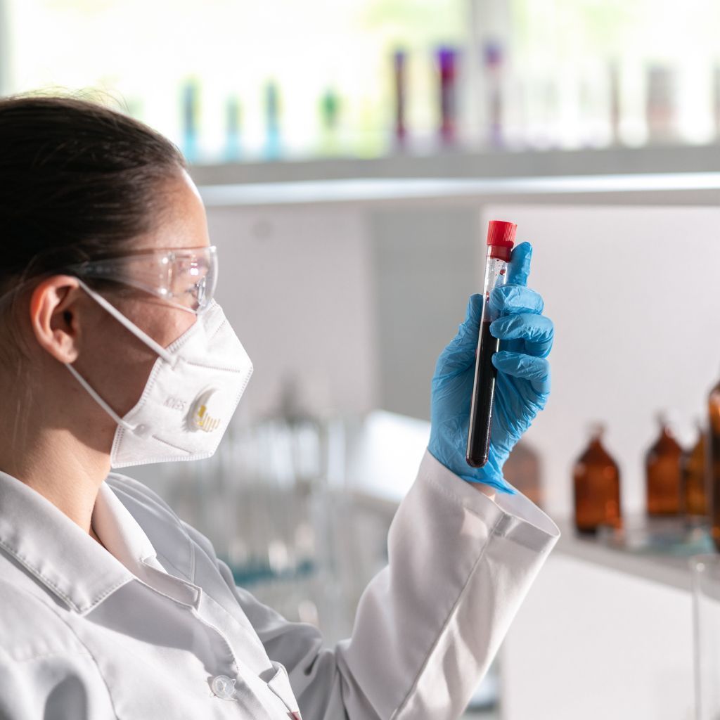 Lab worker in mask and gloves examining blood sample in a lab.
