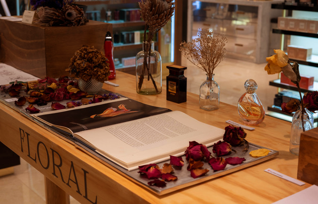 Mesa de madera con un libro abierto, pétalos de flores, jarrones con flores secas y frascos de perfume, dentro de una tienda.