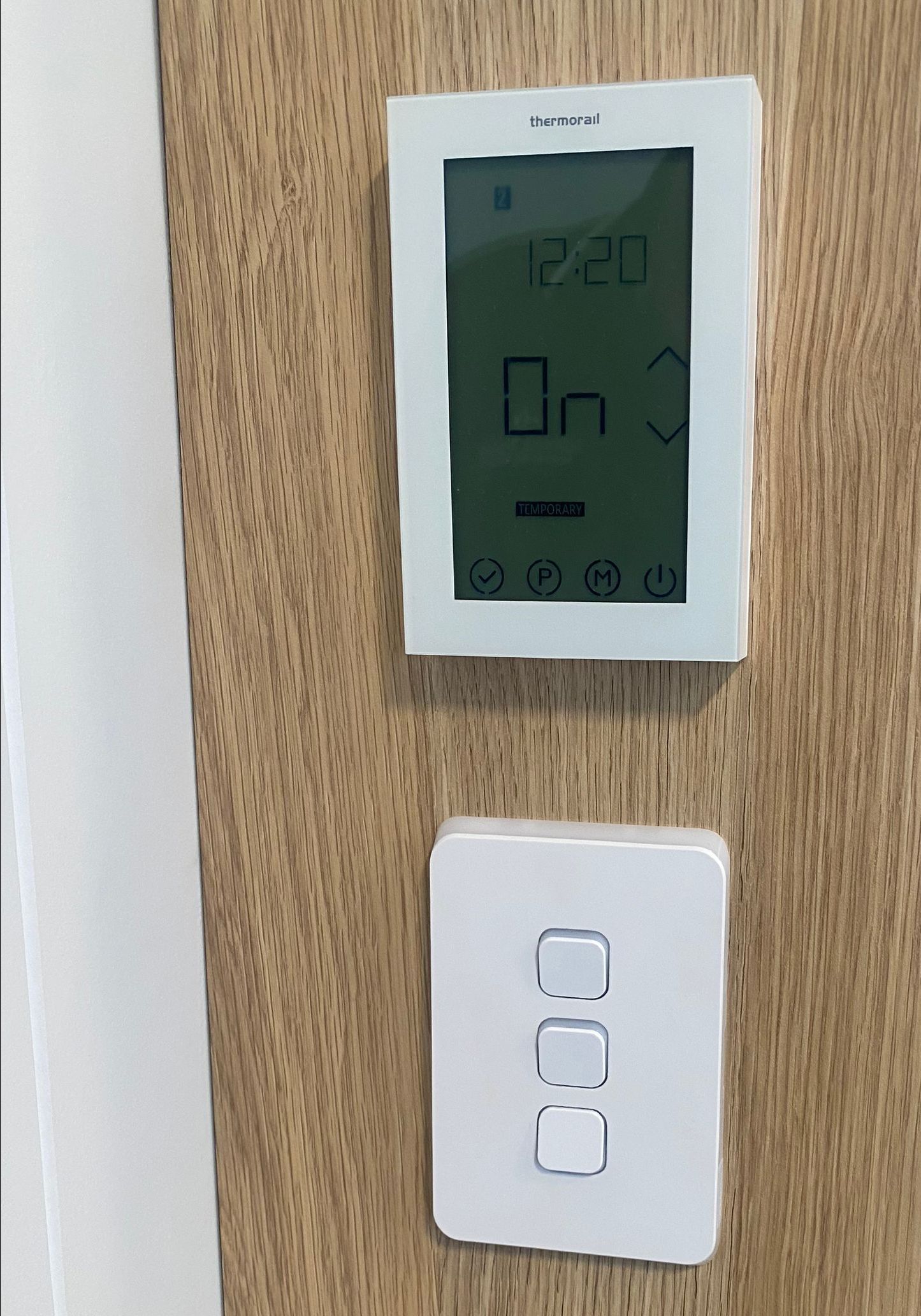 White thermostat and light switch mounted on a wooden wall. Thermostat displays temperature.