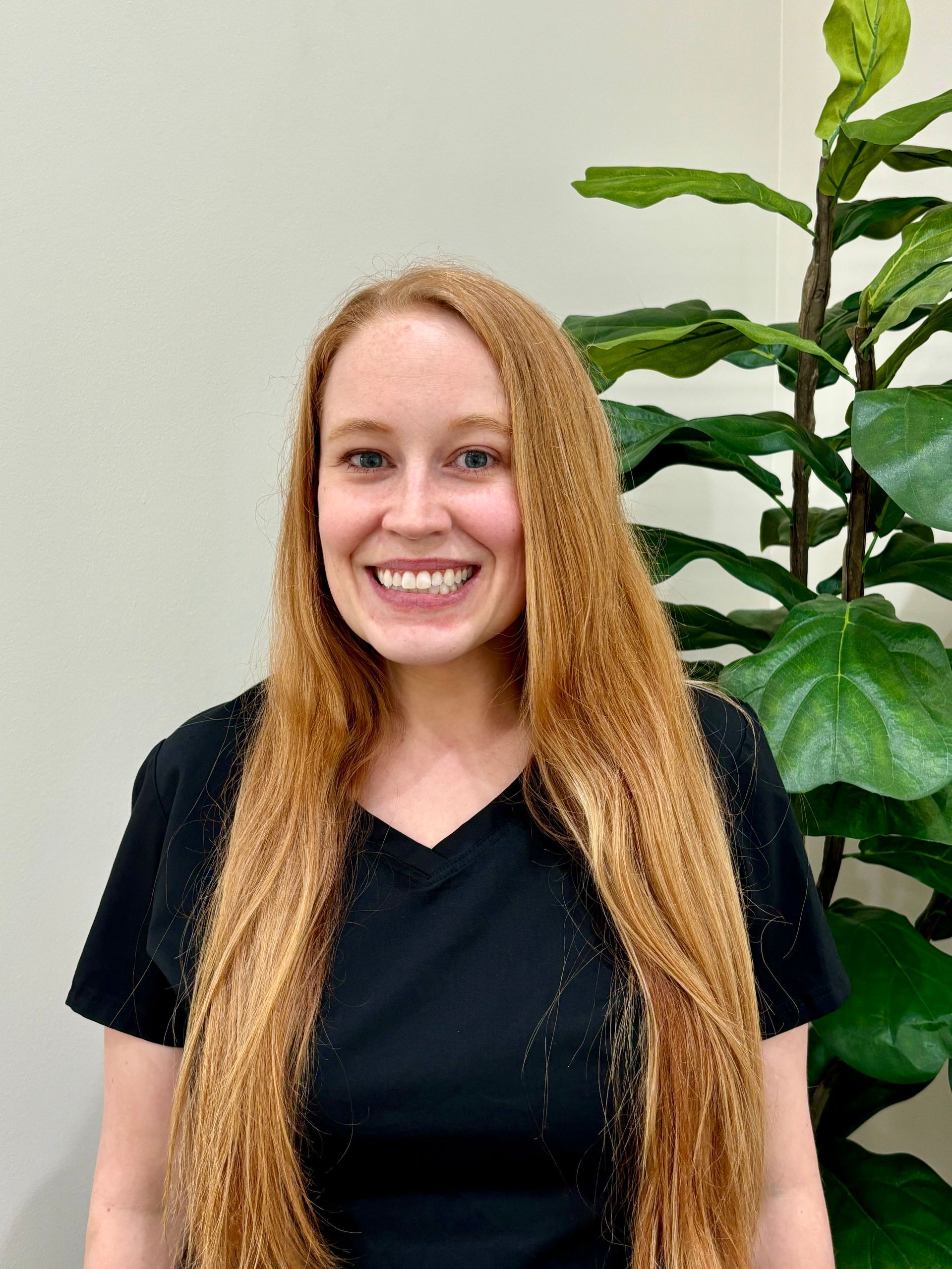 Woman with long red hair smiles, wearing a black shirt, standing by a green plant.
