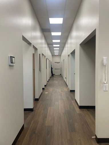 Long, empty hallway with white walls, brown doors, and wood-look flooring.  Fluorescent ceiling lights.