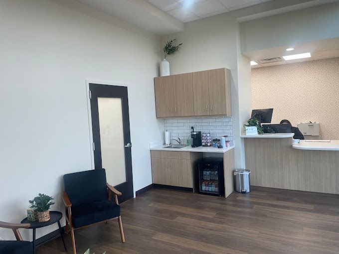 Modern waiting room with a reception desk and coffee station. Includes a black chair, plants, and wooden floors.